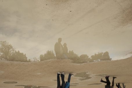 Zwei Personen stehen im Wasserreflexion, umgeben von Bäumen und hellem Himmel.