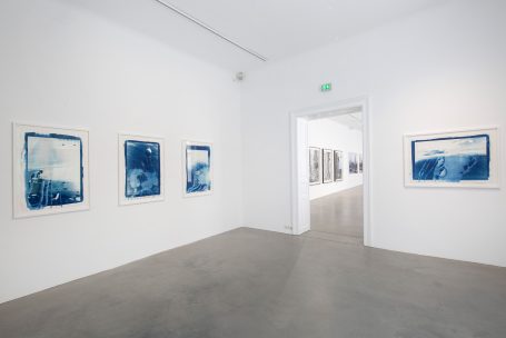 Ausstellungsraum mit weißen Wänden und blauen Kunstwerken in Rahmen.