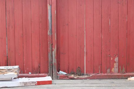 Rote Holzlattenwand mit Beschädigungen und Abfall am Boden.