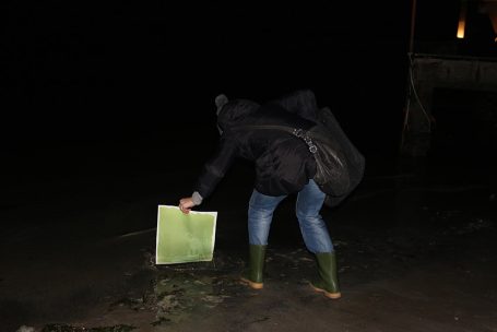 Person in Gummistiefeln beugt sich über den Boden, hält eine grüne Mappe in der Hand.
