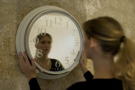 Frau betrachtet ihr Spiegelbild in einer runden Wanduhr.