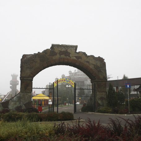 Eingangsbogen zu einem Freizeitpark, unter Nebel und mit bunten Fahrgeschäften im Hintergrund.