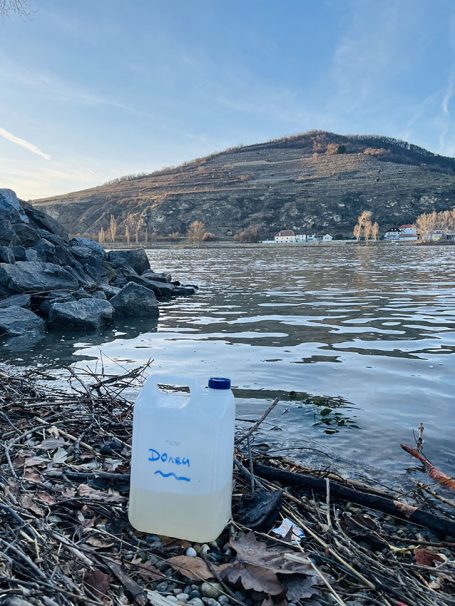 Ein blauer Kanister steht am Ufer eines ruhigen Gewässers, im Hintergrund ein Hügel.