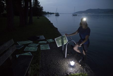 Eine Person mit Stirnlampe steht am Ufer eines ruhigen Gewässers neben Schildern.