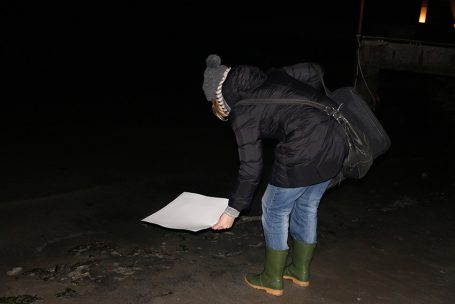 Person mit Mütze und Gummistiefeln beugt sich über ein Blatt Papier am Boden bei Nacht.