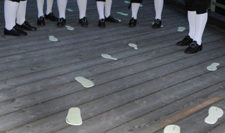 Weiße Fußabdrücke auf einem Holzboden, umgeben von Personen in dunkler Kleidung.
