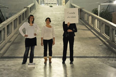 Drei Frauen stehen auf einer Brücke, eine hält ein Schild mit Text.