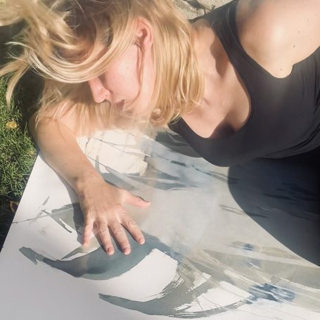 Frau mit blonden Haaren liegt auf einer Leinwand und berührt die Farbe.
