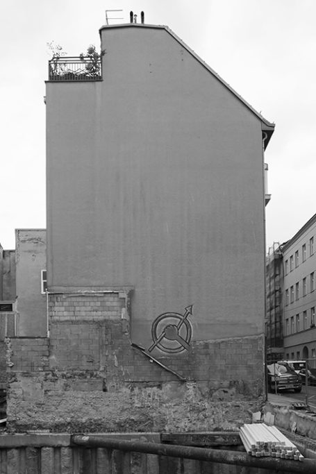 Schmale, graue Gebäudewand mit unvollständiger Putzfläche und Graffiti im Hintergrund.