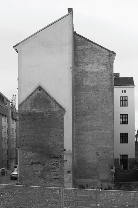 Graue, schlichte Hauswand mit unterschiedlicher Struktur und Form, daneben ein anderes Gebäude.