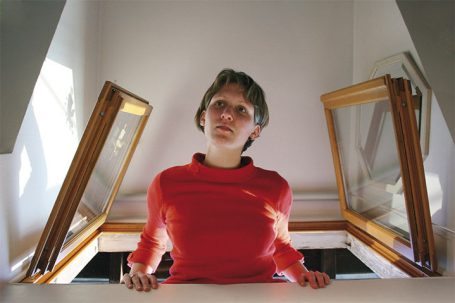 Junge Frau in rotem Pullover lehnt sich aus einem offenen Fenster. Sonnenlicht fällt herein.