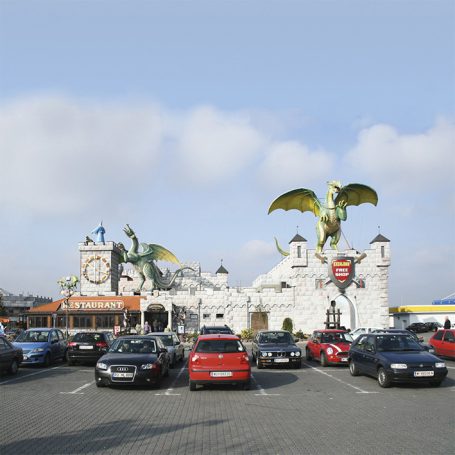 Eingang zu einem Freizeitpark mit Drachen und einer Uhrenfassade, umgeben von Autos.