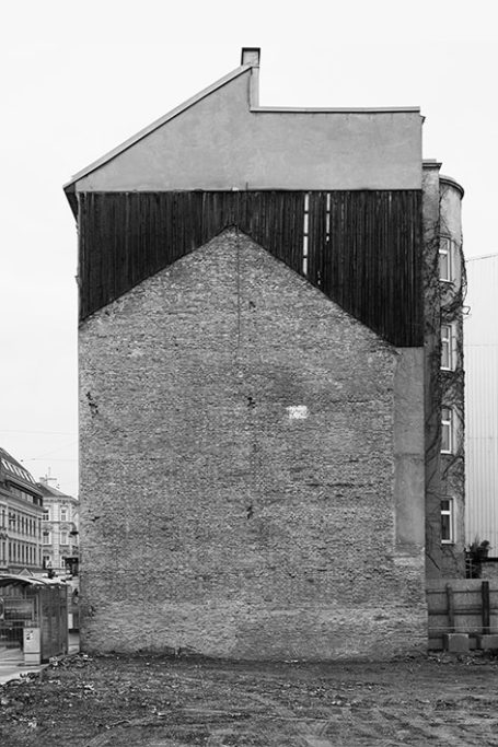 Graue Wand eines Gebäudes mit schieferdachähnlichem Holzabschluss oben.