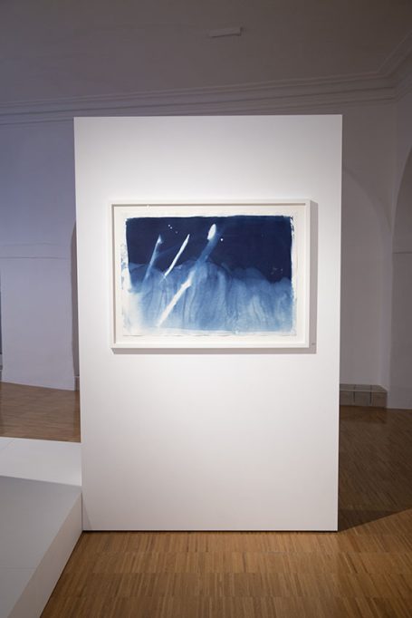 Abstraktes Kunstwerk mit blauen Tönen und weißen Linien auf einer grauen Wand.