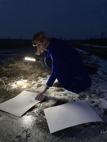 Person in blauer Kleidung kniet im Dunkeln und beleuchtet Papier auf dem Boden.