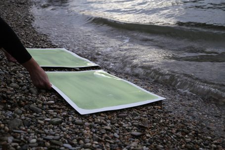 Ein Hand hält zwei grüne Plakate am Ufer, Wellen umspülen den Sand.