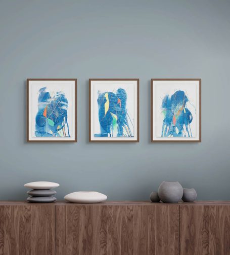Drei gerahmte abstrakte Bilder an einer blauen Wand über einem Holztisch.