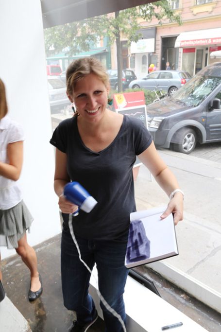 Frau mit Haartrockner in der Hand steht vor einem Fenster mit Blick auf eine Straße.